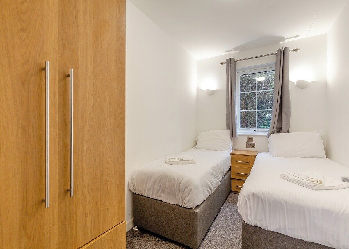 A small, well-lit bedroom with two single beds, a wooden wardrobe, a window with curtains, and a bedside table.