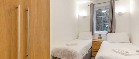 A small, well-lit bedroom with two single beds, a wooden wardrobe, a window with curtains, and a bedside table.