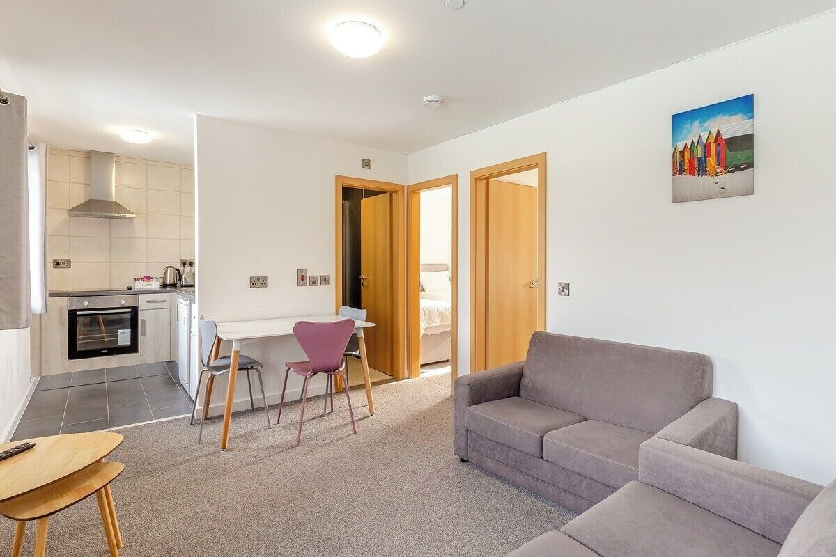 Een moderne woonkamer met een grijze bank, een eethoek met een kleine tafel en stoelen, en een keuken met een fornuis en kasten.
