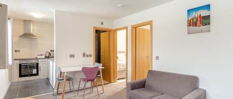 Una sala de estar moderna con un sofá gris, un comedor con una mesa pequeña y sillas, y una cocina con estufa y armarios.