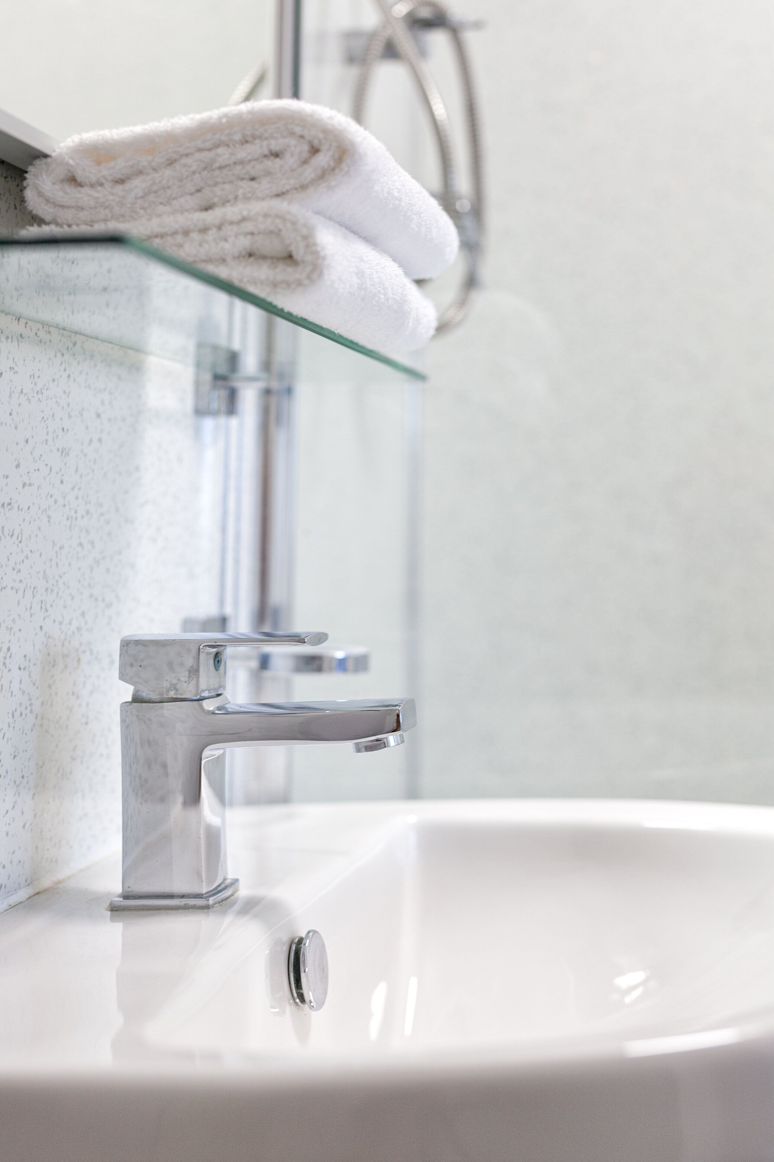 A bathroom with a glass shelf holding a rolled white towel, a chrome faucet, and a white sink.