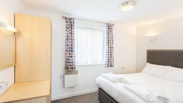 A hotel room with a bed, a bedside table, a wardrobe, a window with curtains, and a wall-mounted light fixture.