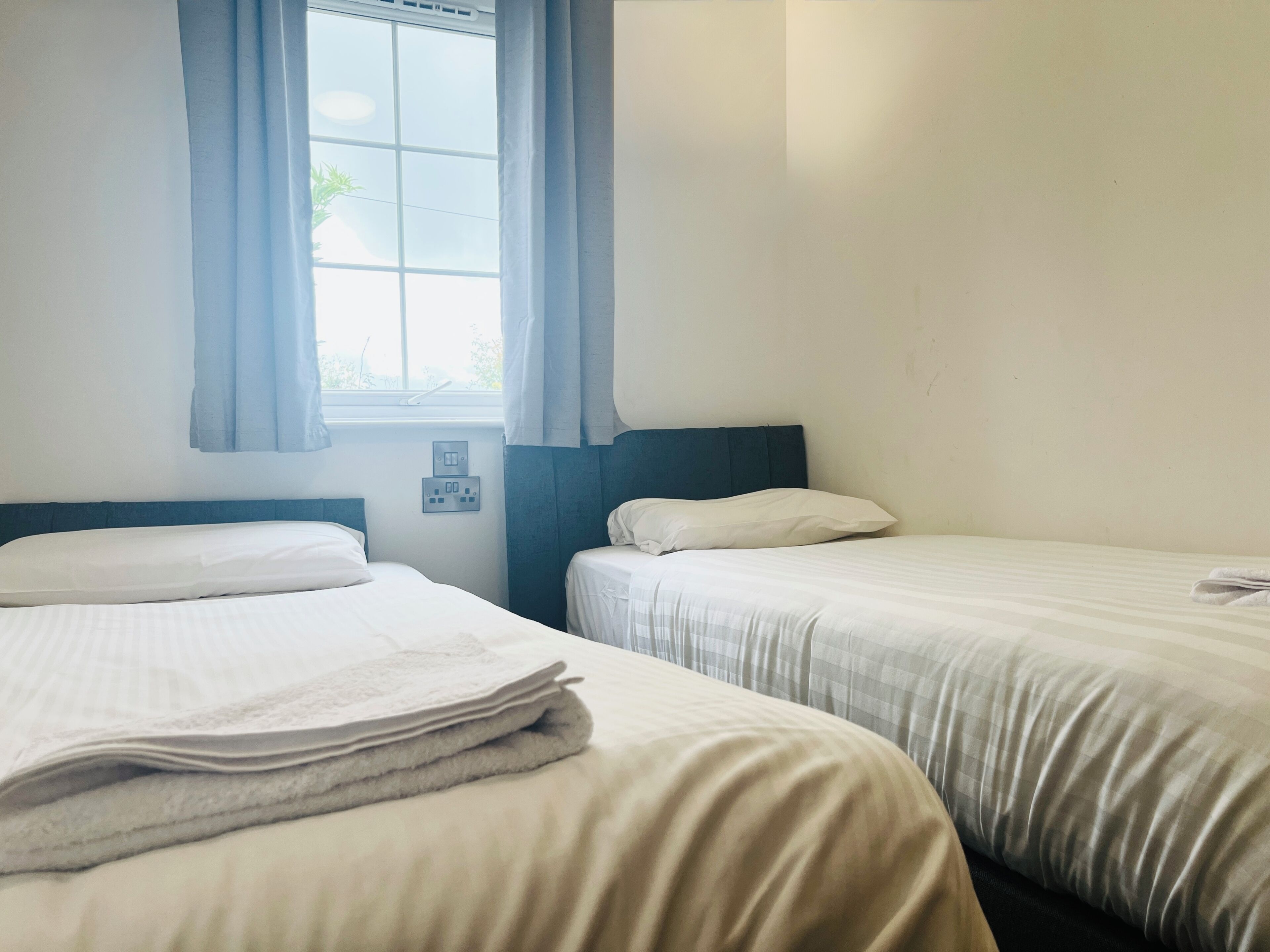 A room with two beds, a window with curtains, and a wall socket.