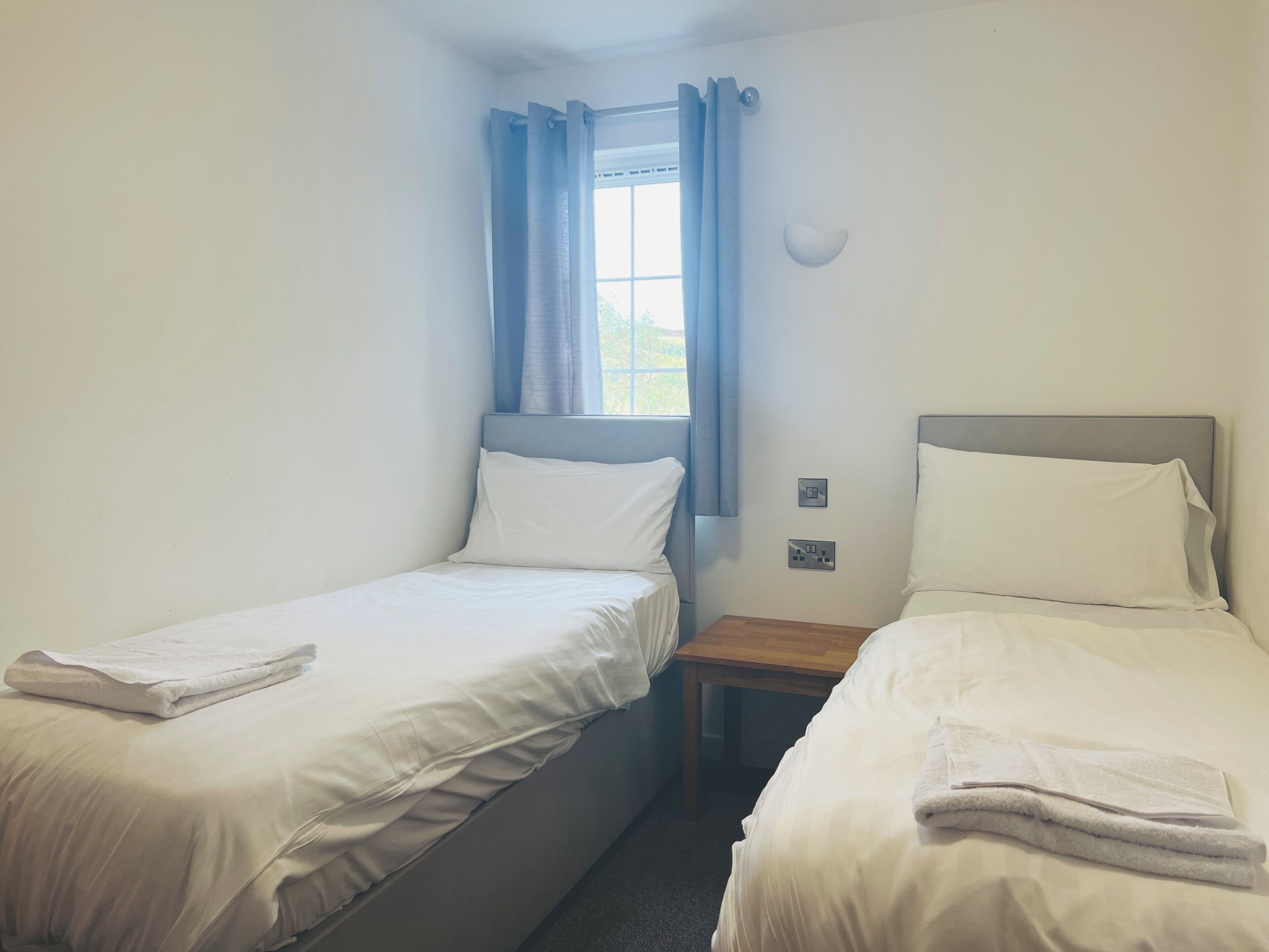 Habitación de hotel con dos camas individuales, una ventana con cortinas, una mesita de noche y una lámpara de pared.
