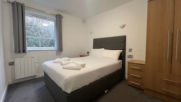 Un dormitorio con cama, ventana con cortinas, radiador, armario de madera y mesita de noche.