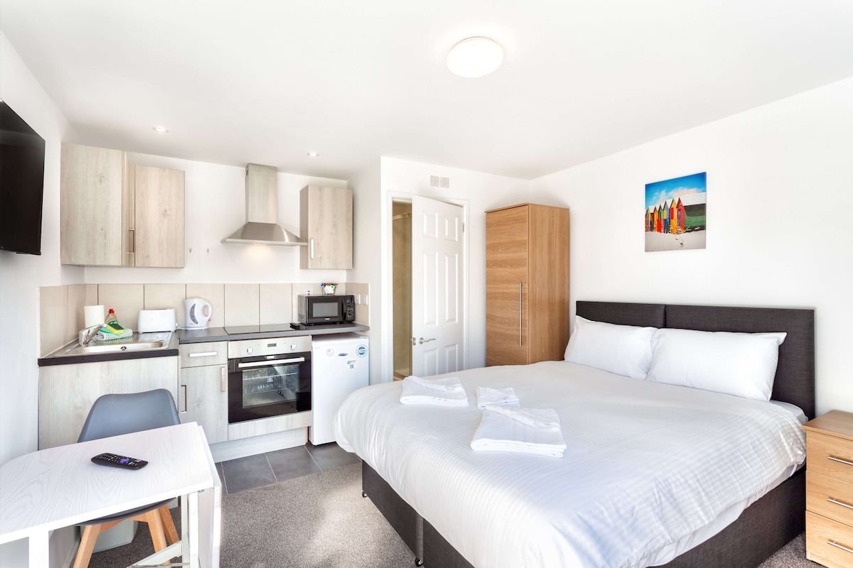 Een moderne hotelkamer met kitchenette, een bed met netjes gevouwen handdoeken, een nachtkastje en een schilderij aan de muur.