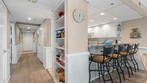 A hallway with a kitchen area, featuring white cabinetry, a clock, and a bar seating area with chairs.