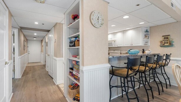 A hallway with a kitchen area, featuring white cabinetry, a clock, and a bar seating area with chairs. - Rainbow - Gorgeous 3 Bedroom Condo with Ocean Views and Outdoor Pool! (Ocean City)