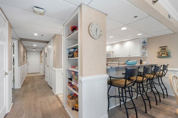 A hallway with a kitchen area, featuring white cabinetry, a clock, and a bar seating area with chairs.