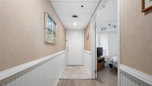 A hallway with a door leading to a bedroom. The hallway has a white baseboard, a painting on the wall, and a ceiling light. - Rainbow - Gorgeous 3 Bedroom Condo with Ocean Views and Outdoor Pool! (Ocean City)