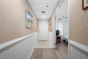 Un pasillo con una puerta que da a un dormitorio. El pasillo tiene un zócalo blanco, un cuadro en la pared y una lámpara en el techo.