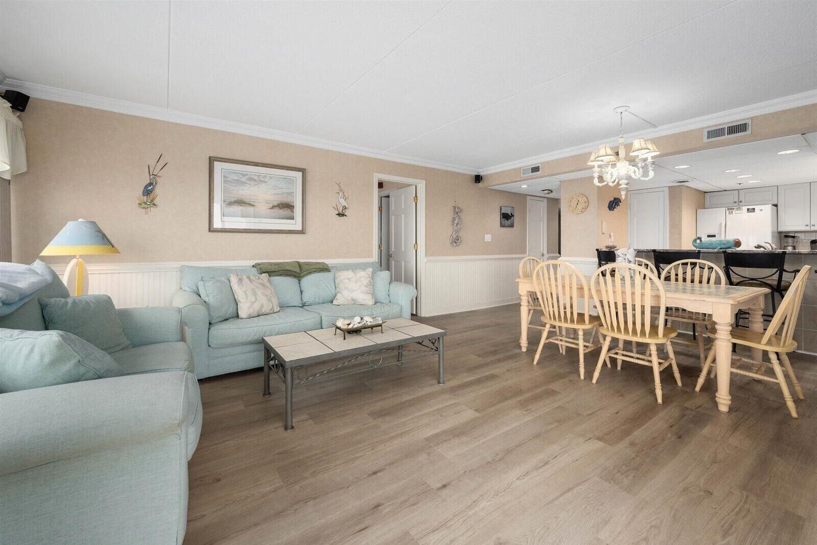 Ein Wohnzimmer mit einer Couch, einem Couchtisch und einem Esstisch mit Stühlen. Eine Lampe, ein Gemälde an der Wand und eine Küchenzeile im Hintergrund sind ebenfalls zu sehen.