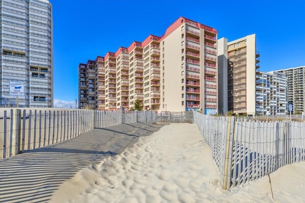 Un hotel frente a la playa con varios edificios altos, un paseo marítimo de arena y una valla de madera.