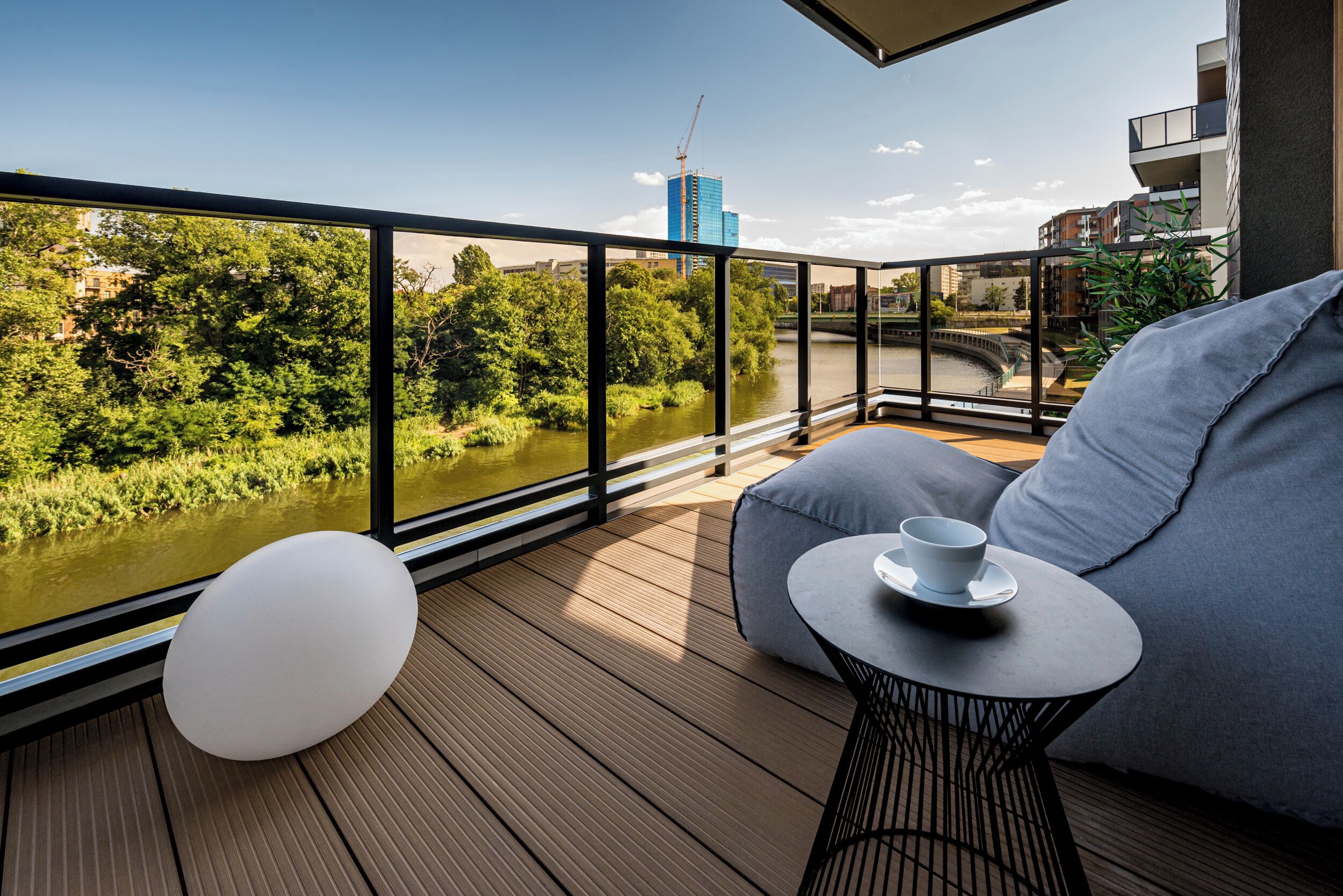 A balcony with a view of a river, a modern building in the background, and a seating area with a table and a cup.