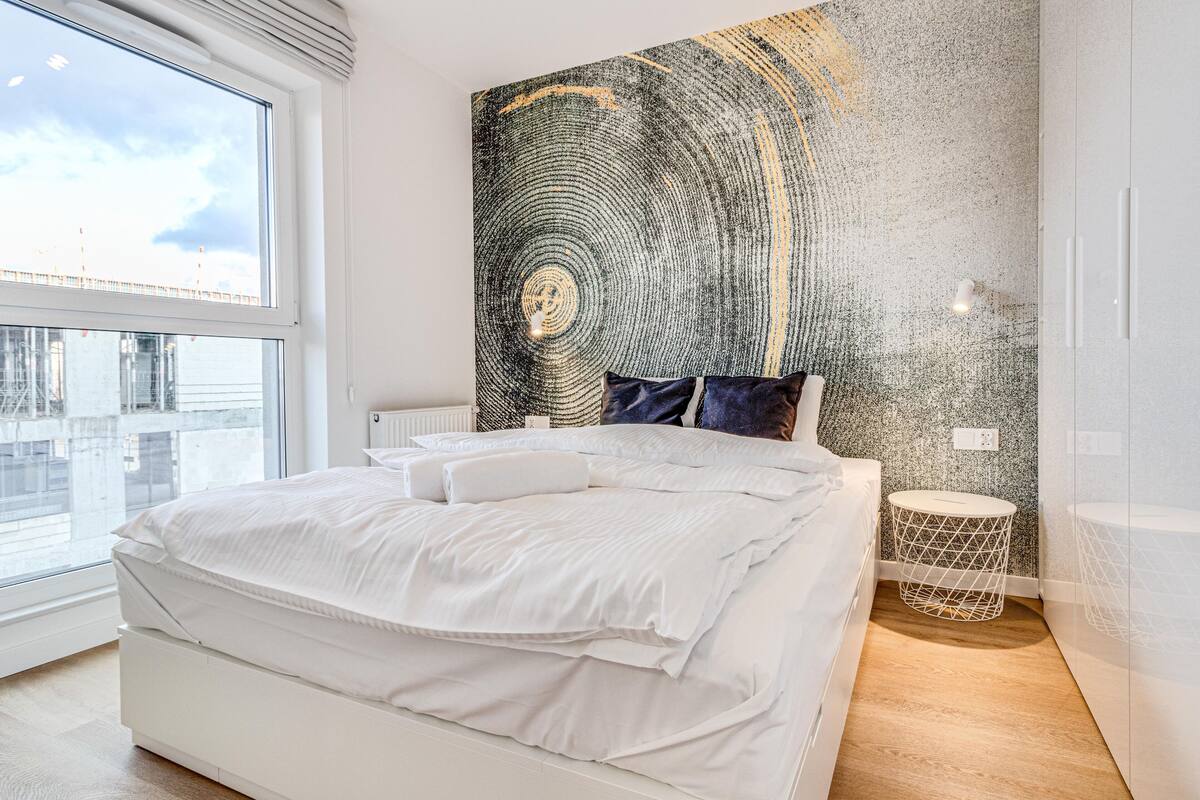A modern bedroom with a large bed, a city view, and a textured wall art piece.