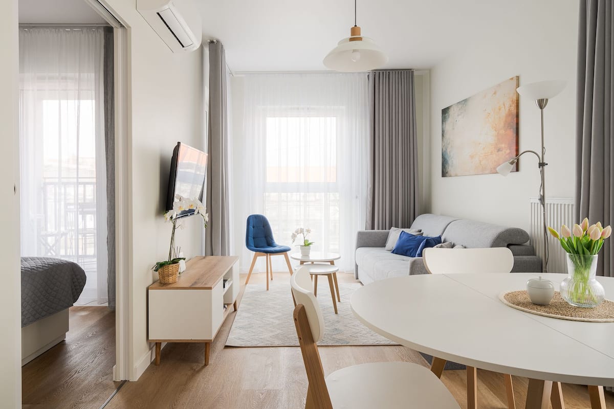 A modern hotel room with a bed, a desk, a dining table, a sofa, and a TV.