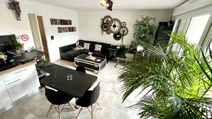 A modern living room with a black sofa, a dining table with chairs, a kitchen area with a countertop, and a large potted plant.