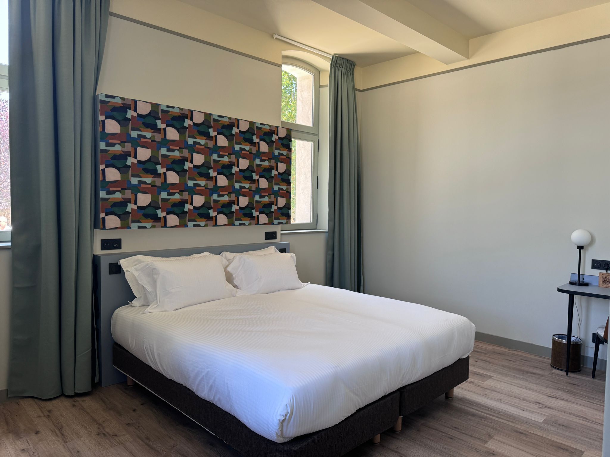 A modern bedroom with a large bed, grey curtains, a geometric wall art piece, and a small bedside table with a lamp.