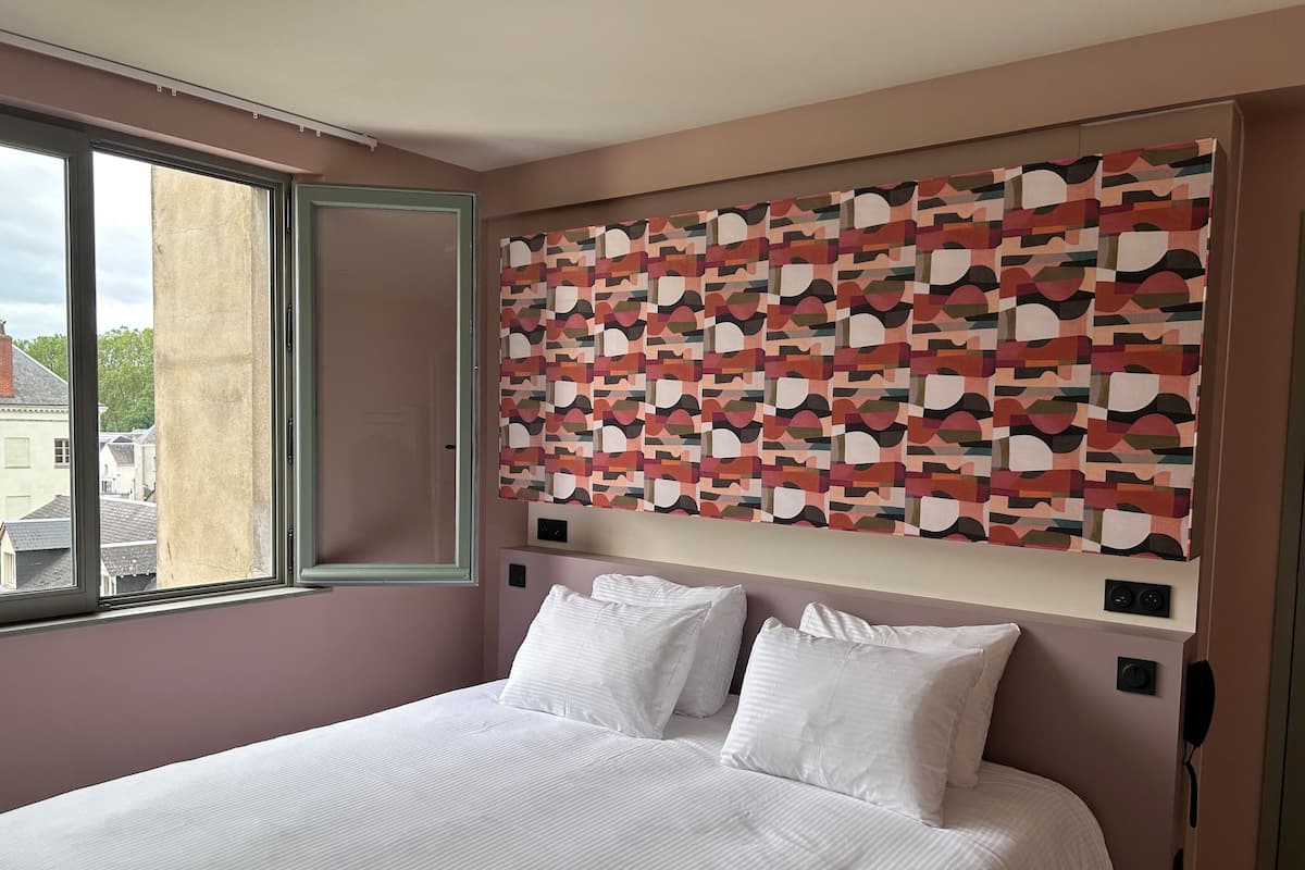 A hotel room with a large bed, a window with a view of buildings, and a colorful abstract wall art.