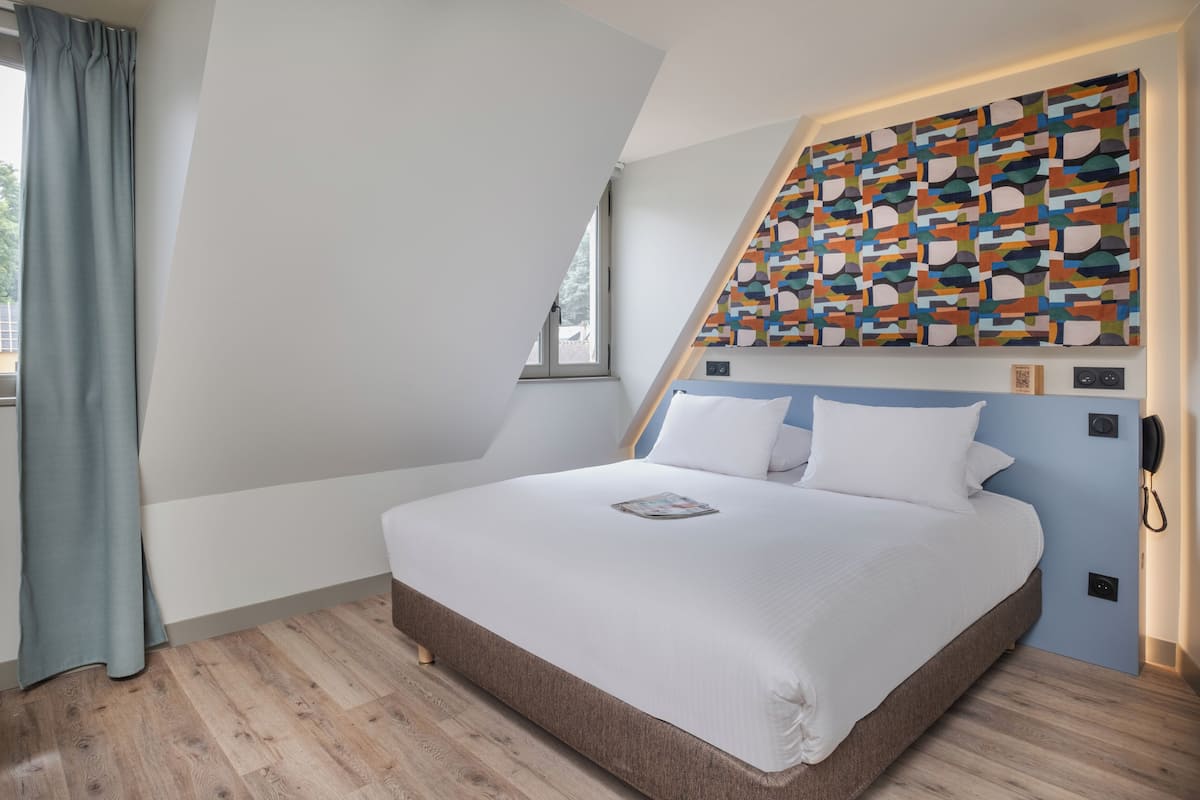 A modern bedroom with a sloped ceiling, a large bed with white bedding, a colorful abstract wall art, and a window with curtains.