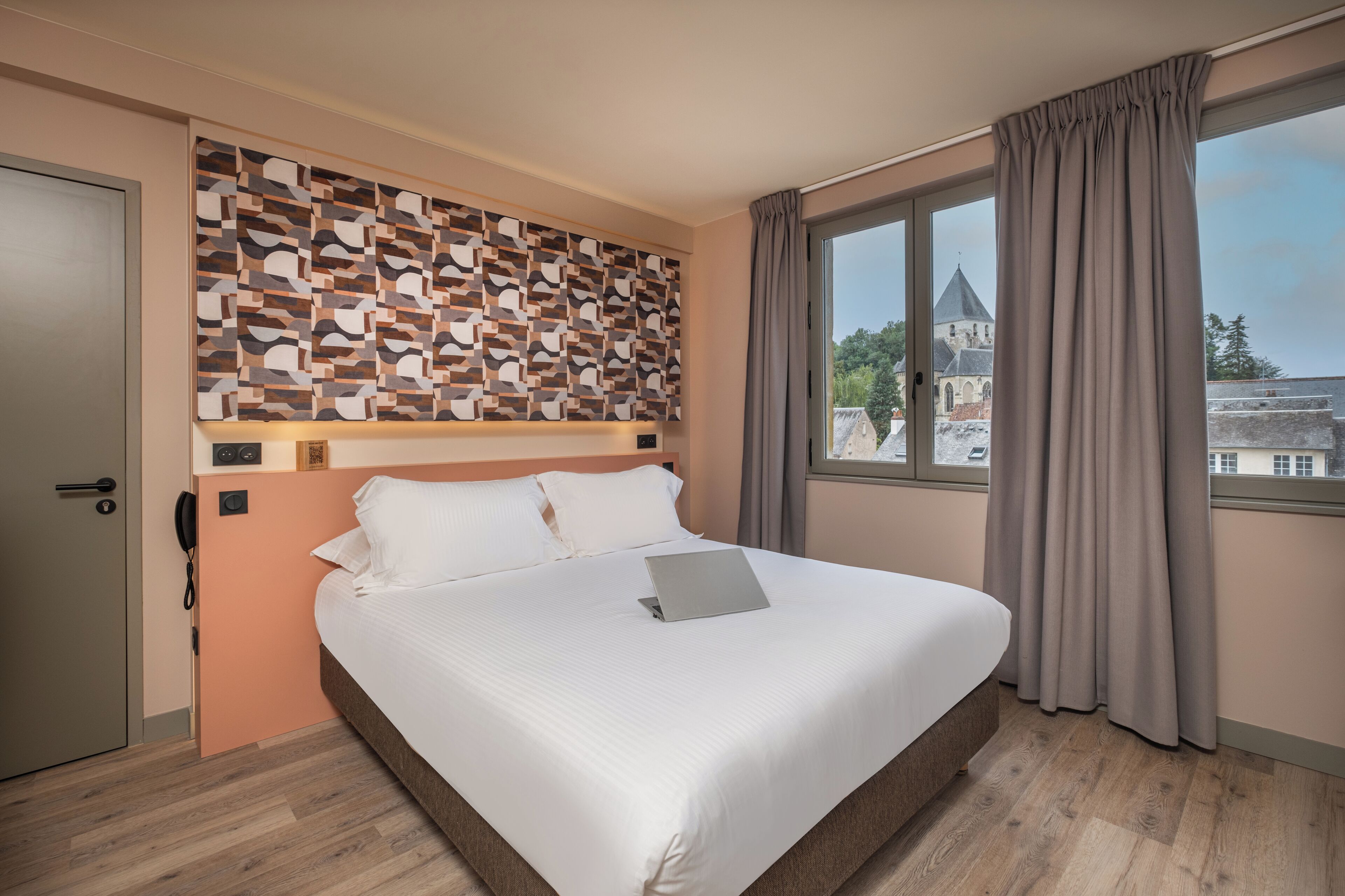 A hotel room with a bed, a patterned wall, a window with curtains, and a view of a building.