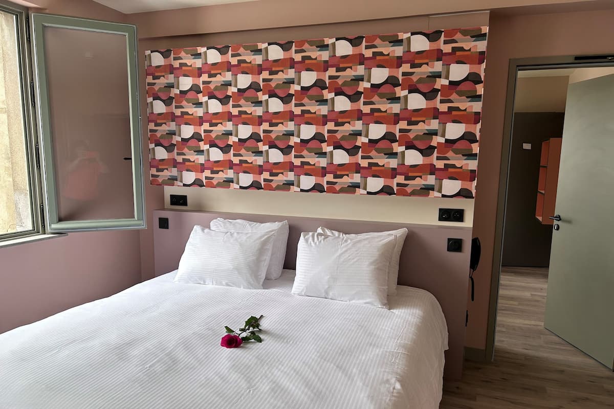 A hotel room with a bed, white bedding, a decorative wall panel, and a door leading to another room.