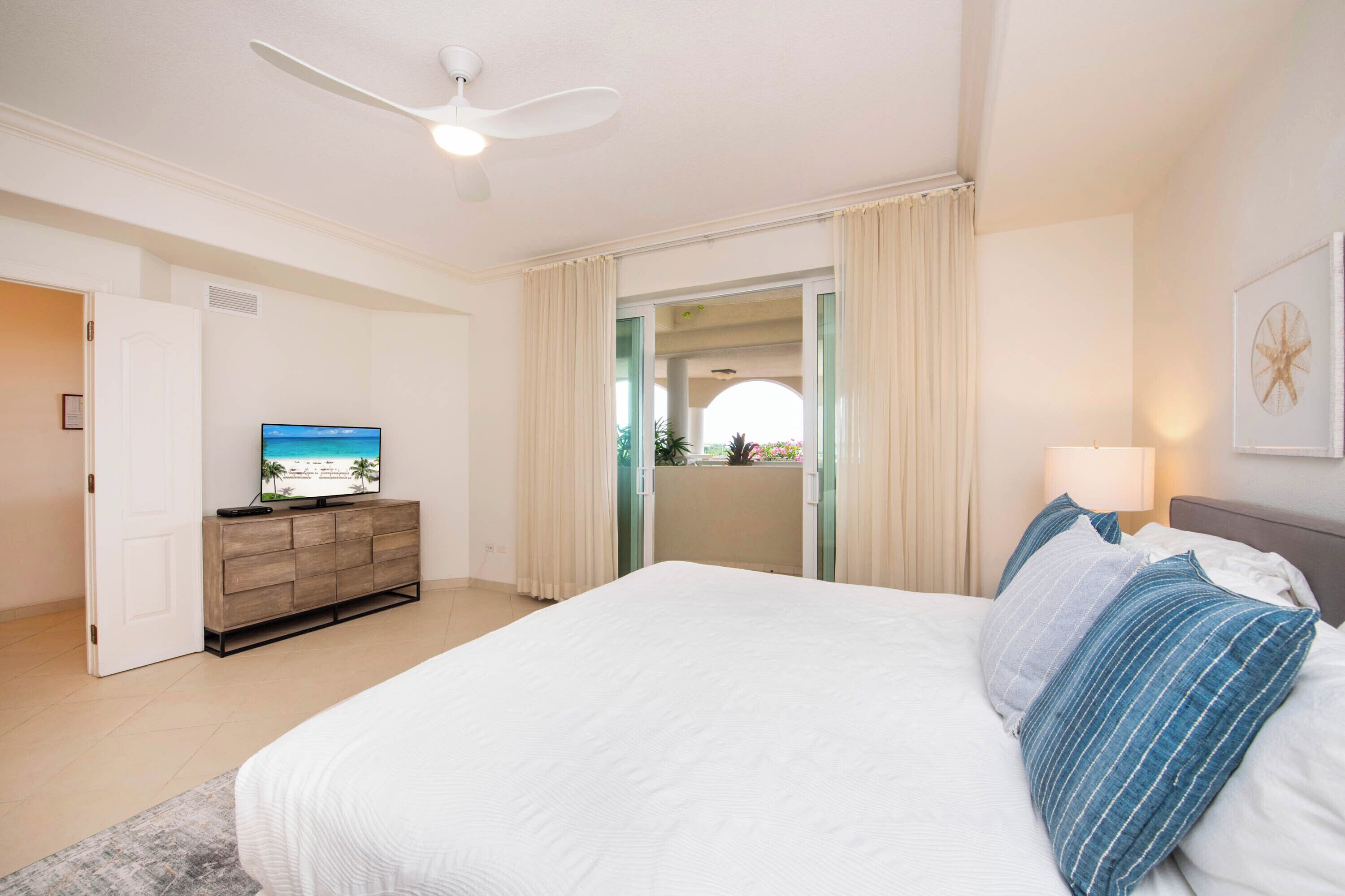 A bedroom with a bed, a TV on a wooden dresser, a ceiling fan, and a view through a door.