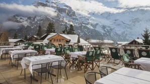 A restaurant with outdoor seating overlooking a snow-covered mountain. Tables are set with white tablecloths and green chairs. - Drei Berge Hotel (Mürren)