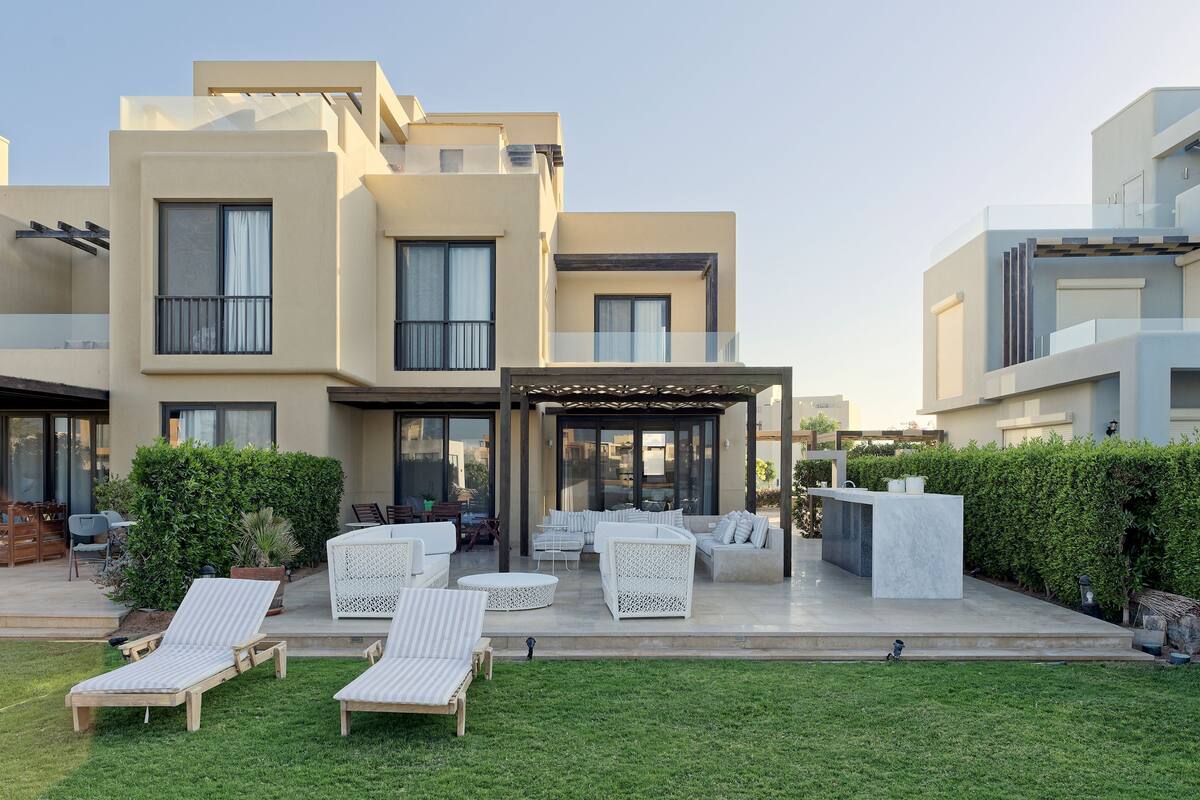 A modern multi-story building with a covered outdoor seating area, lounge chairs, and a well-maintained lawn.
