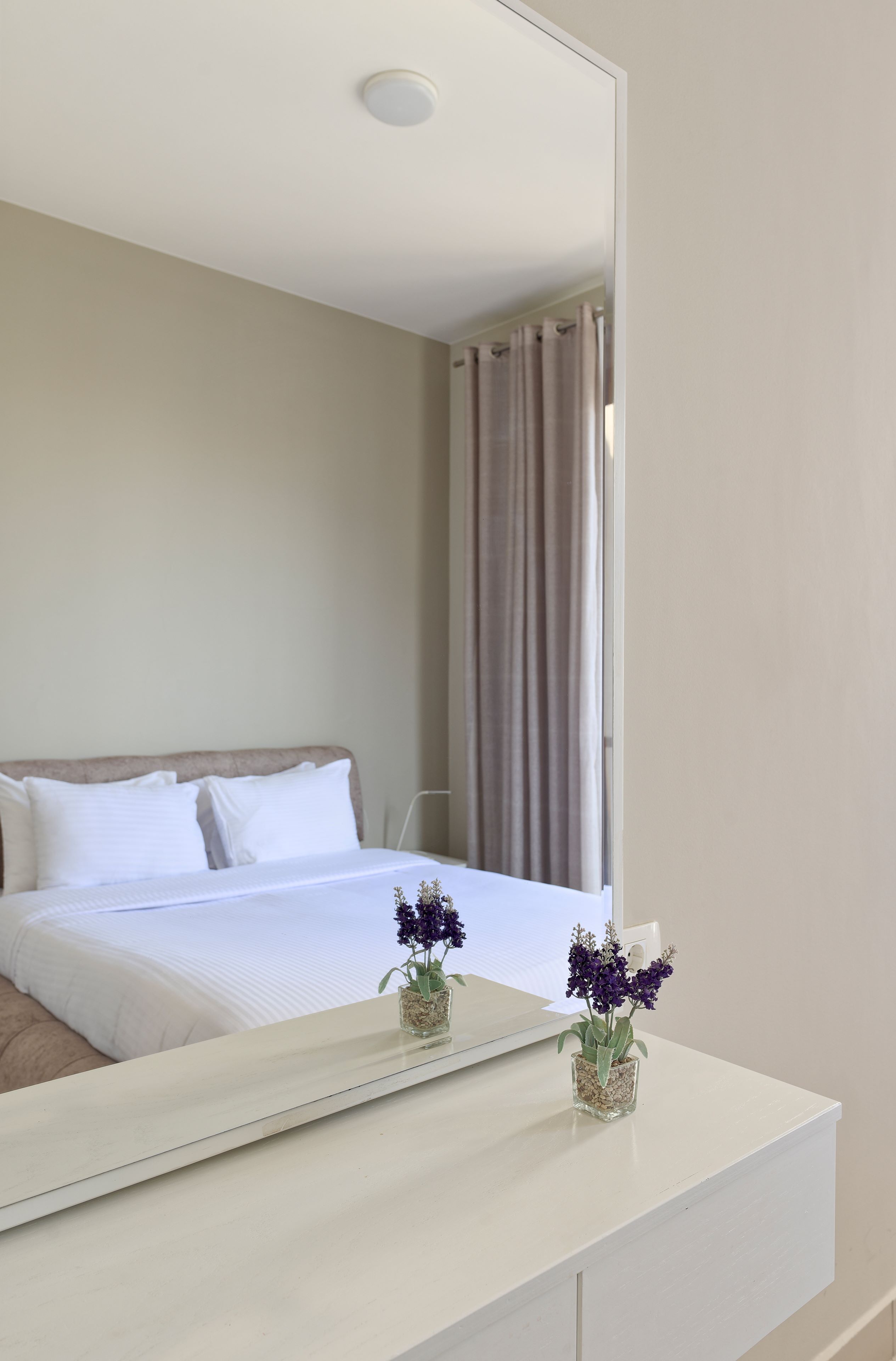 A hotel room with a bed, a white bedside table, a small vase with flowers, and a mirror reflecting the room.