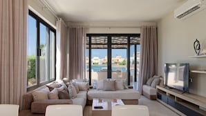 Un moderno salón con un sofá grande, una mesa de centro y un televisor. Amplias ventanas con cortinas ofrecen vistas a una piscina y edificios.