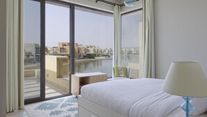 Habitación de hotel con un amplio ventanal corredero que da a un balcón, una cama con edredón blanco, una mesita de noche y una lámpara.