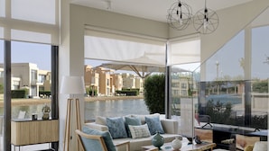 Una sala de estar moderna con sofá, mesa de centro y lámpara de pie. Se ve un ventanal con vistas a edificios y una masa de agua.