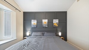 Une chambre avec un grand lit, deux tables de chevet avec des lampes, une fenêtre avec des stores et trois œuvres d’art accrochées au mur, représentant des tournesols.