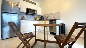 Een compacte keuken met een koelkast, magnetron, spoelbak en een kleine eettafel met twee houten stoelen.