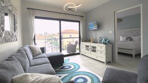 Een moderne woonkamer met een grijze bank, een kleine salontafel en een televisie op een standaard. Er is een balkon met uitzicht op de omgeving.