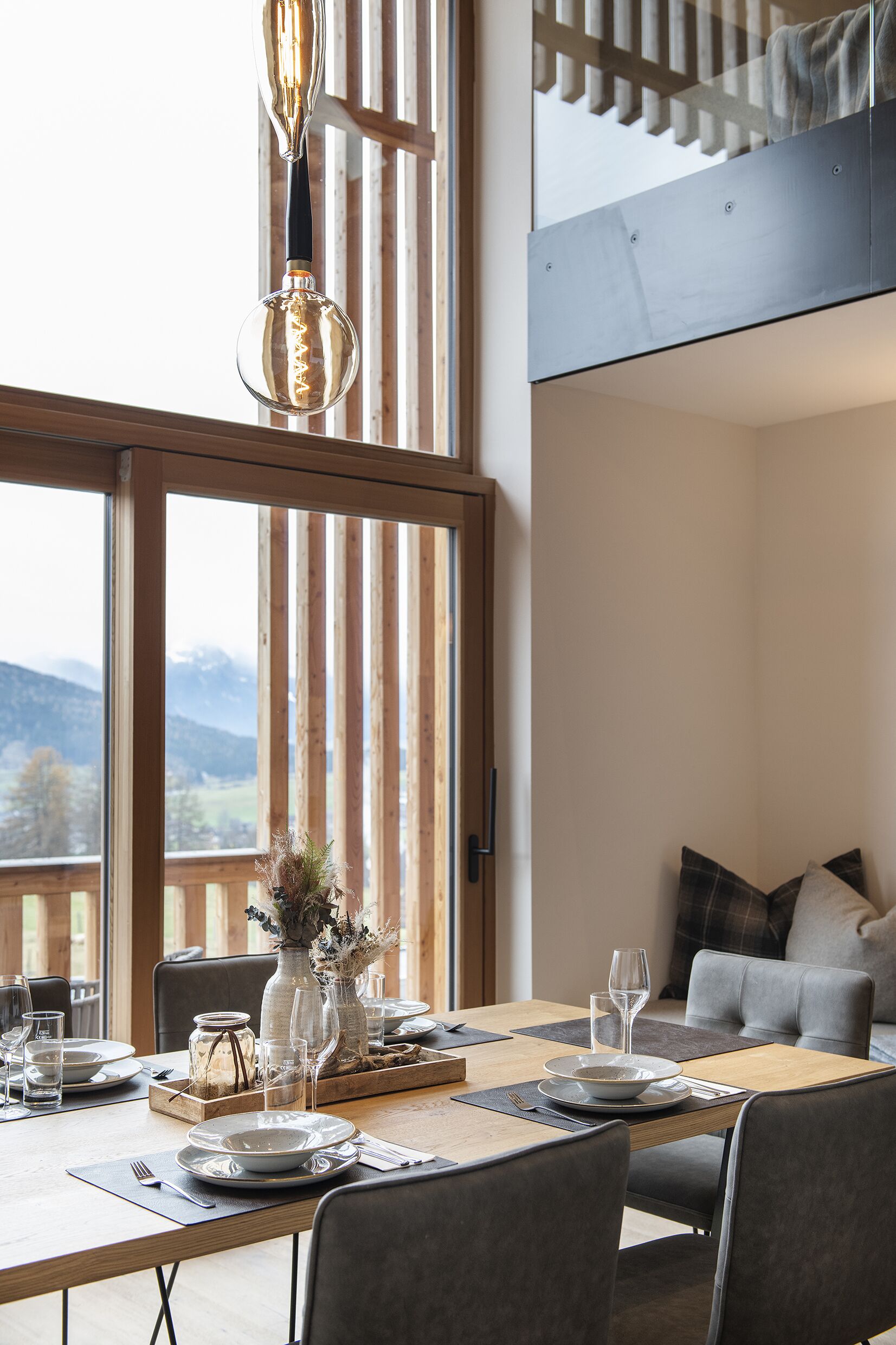 Zona pranzo con tavolo in legno apparecchiato per due persone, un vaso con fiori e vista sulle montagne attraverso ampie finestre.