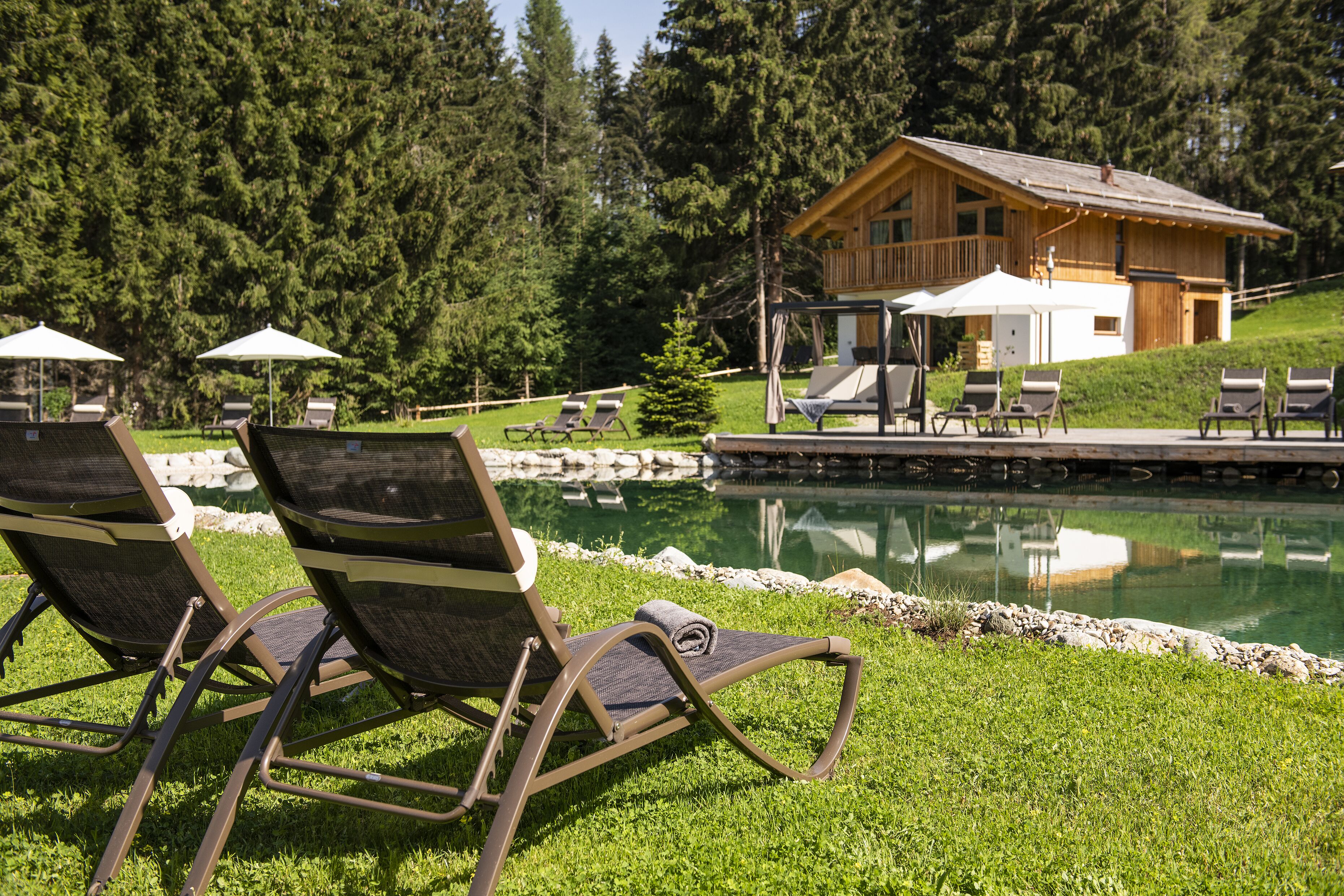 A poolside area with lounge chairs and umbrellas, a wooden cabin, and a reflection in the water.