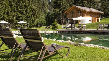 A poolside area with lounge chairs and umbrellas, a wooden cabin, and a reflection in the water.