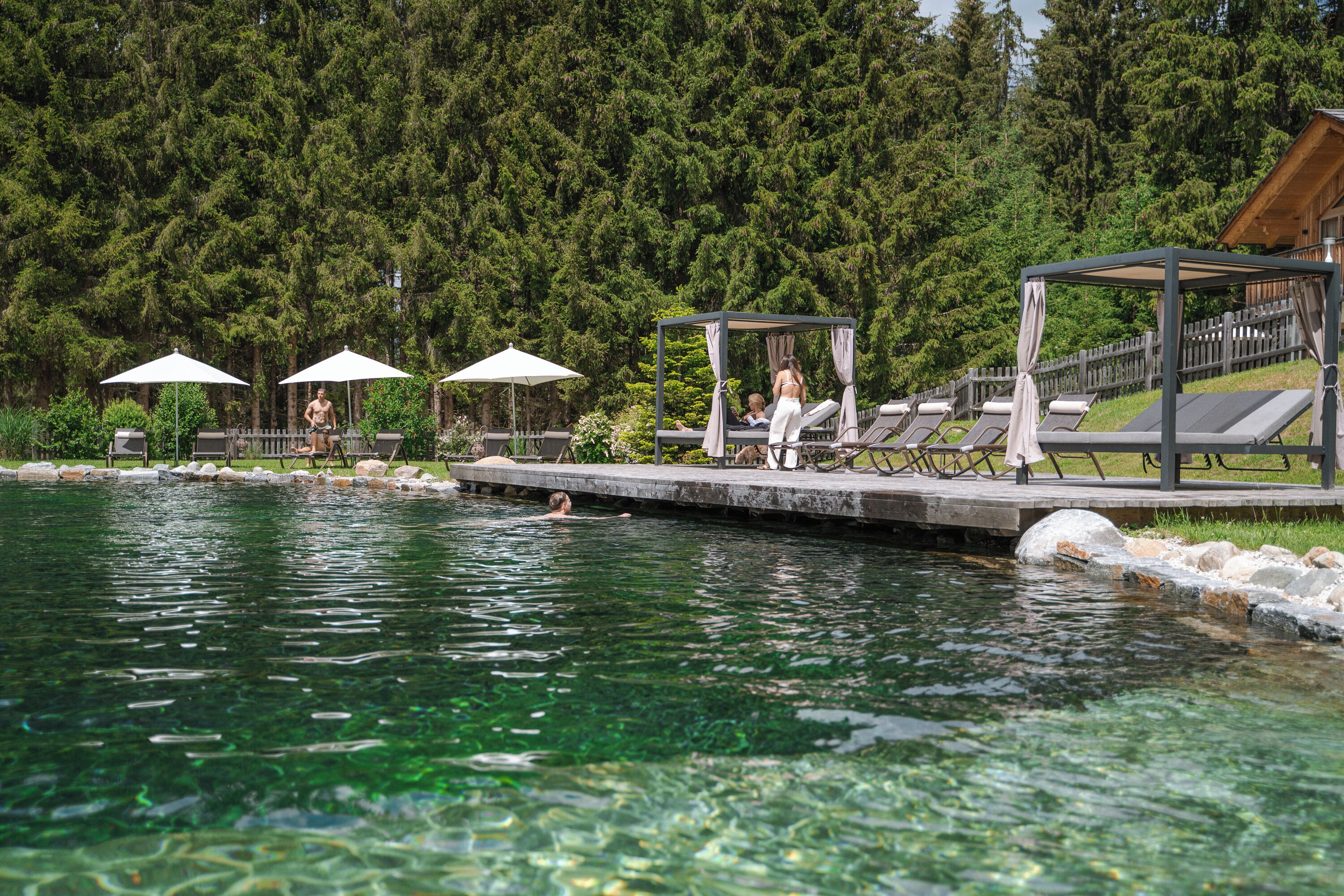 A swimming pool with lounge chairs and umbrellas, surrounded by a wooden deck and a forested area.