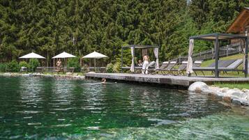 A swimming pool with lounge chairs and umbrellas, surrounded by a wooden deck and a forested area.