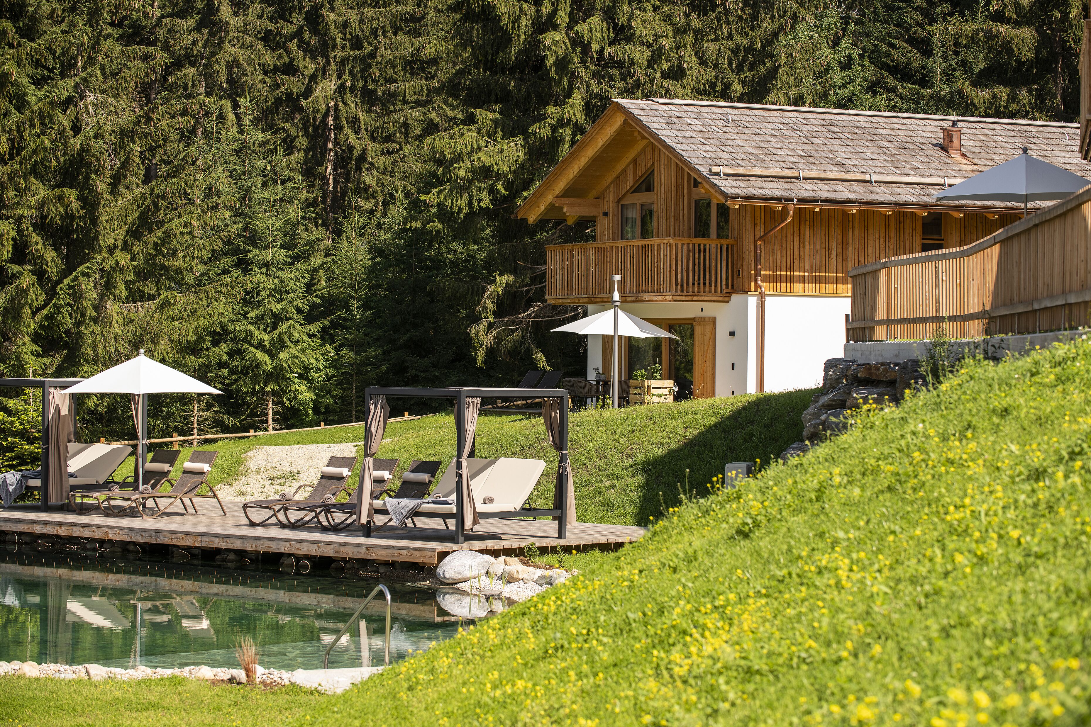 A wooden cabin with a deck and lounge chairs by a pool, surrounded by greenery and trees.