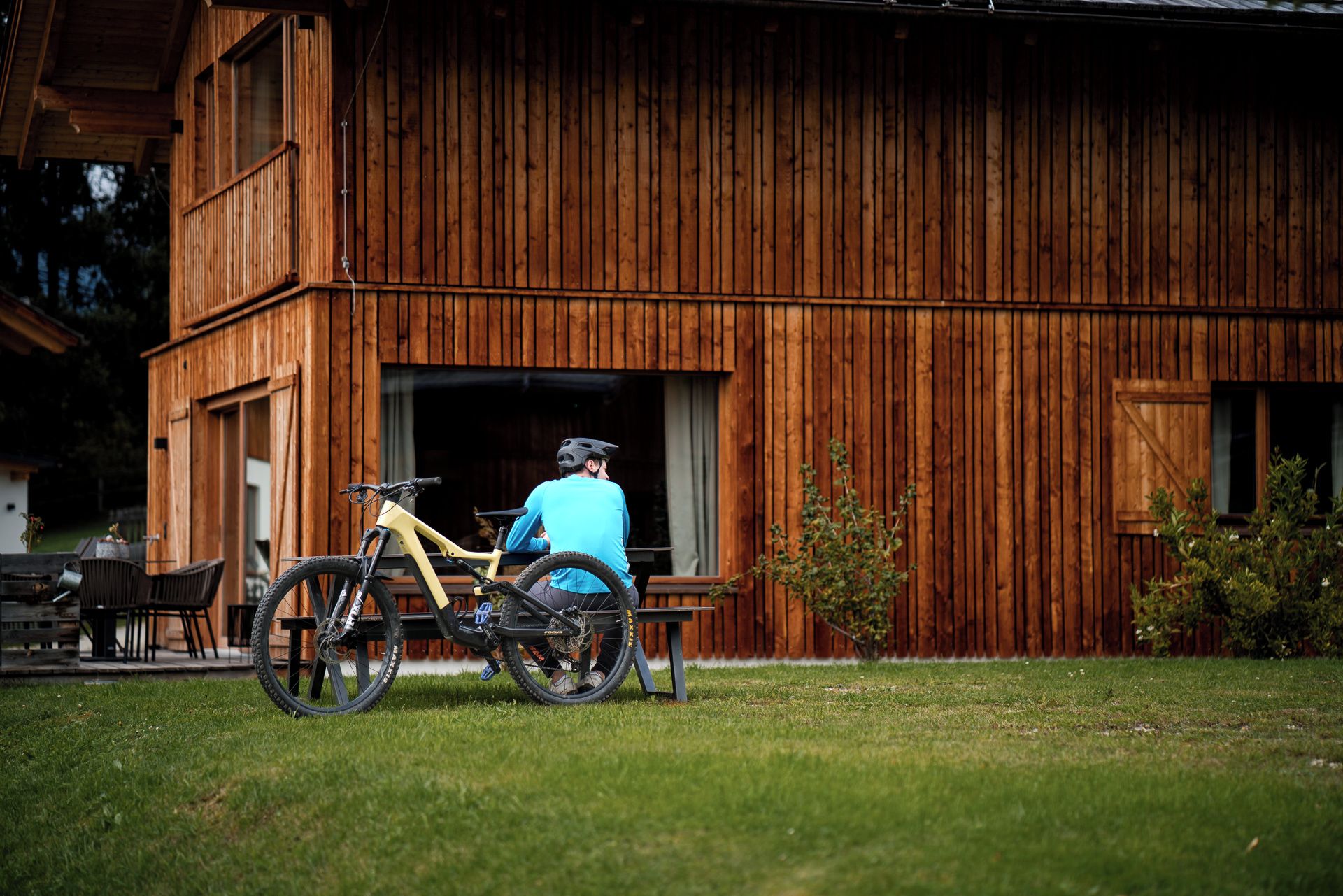Ein Holzhaus mit großem offenen Durchgang, einem Fahrrad davor und einer Person, die auf einer Bank sitzt.