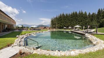 Una piscina cristalina rodeada de un borde de piedra, con tumbonas y sombrillas, con árboles y montañas como fondo.