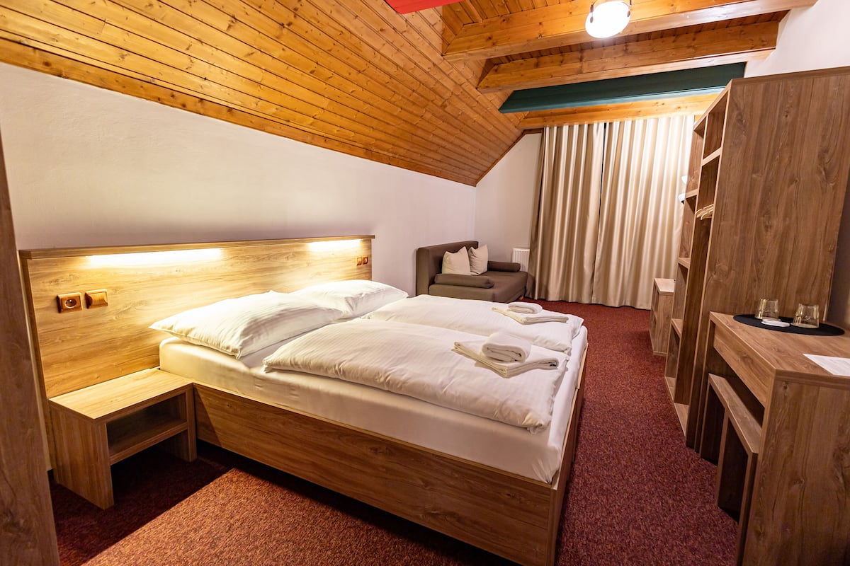 A wooden bed with white linens, a bedside table, a chair, and a wooden wardrobe in a room with a sloped ceiling and a red carpet.