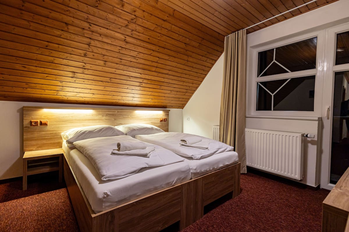A bedroom with a wooden ceiling, a bed with white linens, a nightstand, a window with curtains, and a radiator.