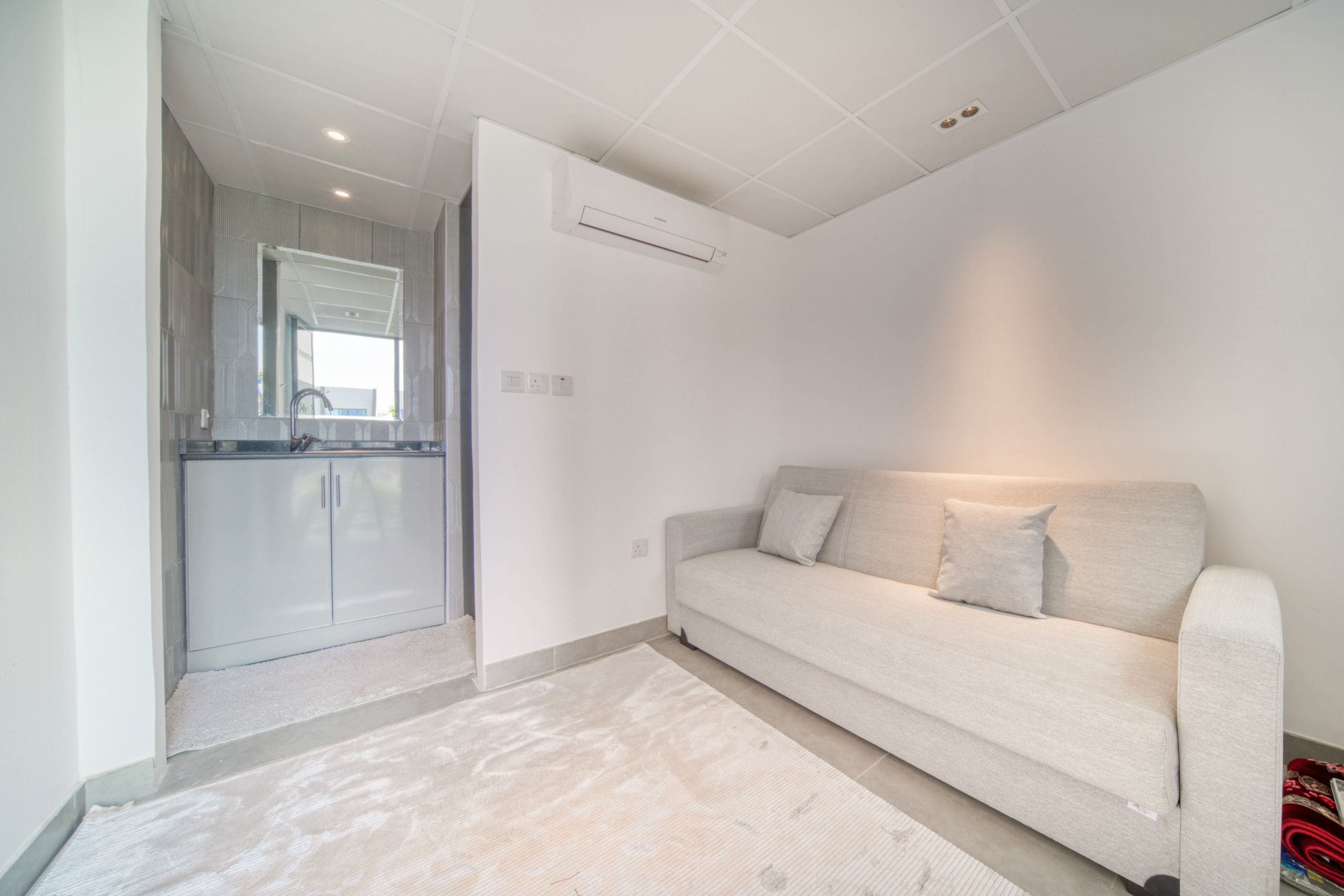 A modern living room with a sofa, a kitchen area visible through an open door, and a ceiling with recessed lighting.