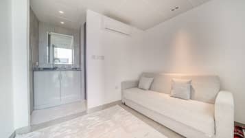 A modern living room with a sofa, a kitchen area visible through an open door, and a ceiling with recessed lighting.