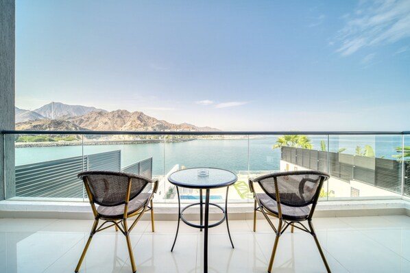 A balcony with two wicker chairs and a small round table, overlooking a coastal area with mountains and a body of water.
