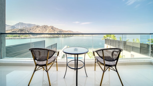 A balcony with two wicker chairs and a small round table, overlooking a coastal area with mountains and a body of water.